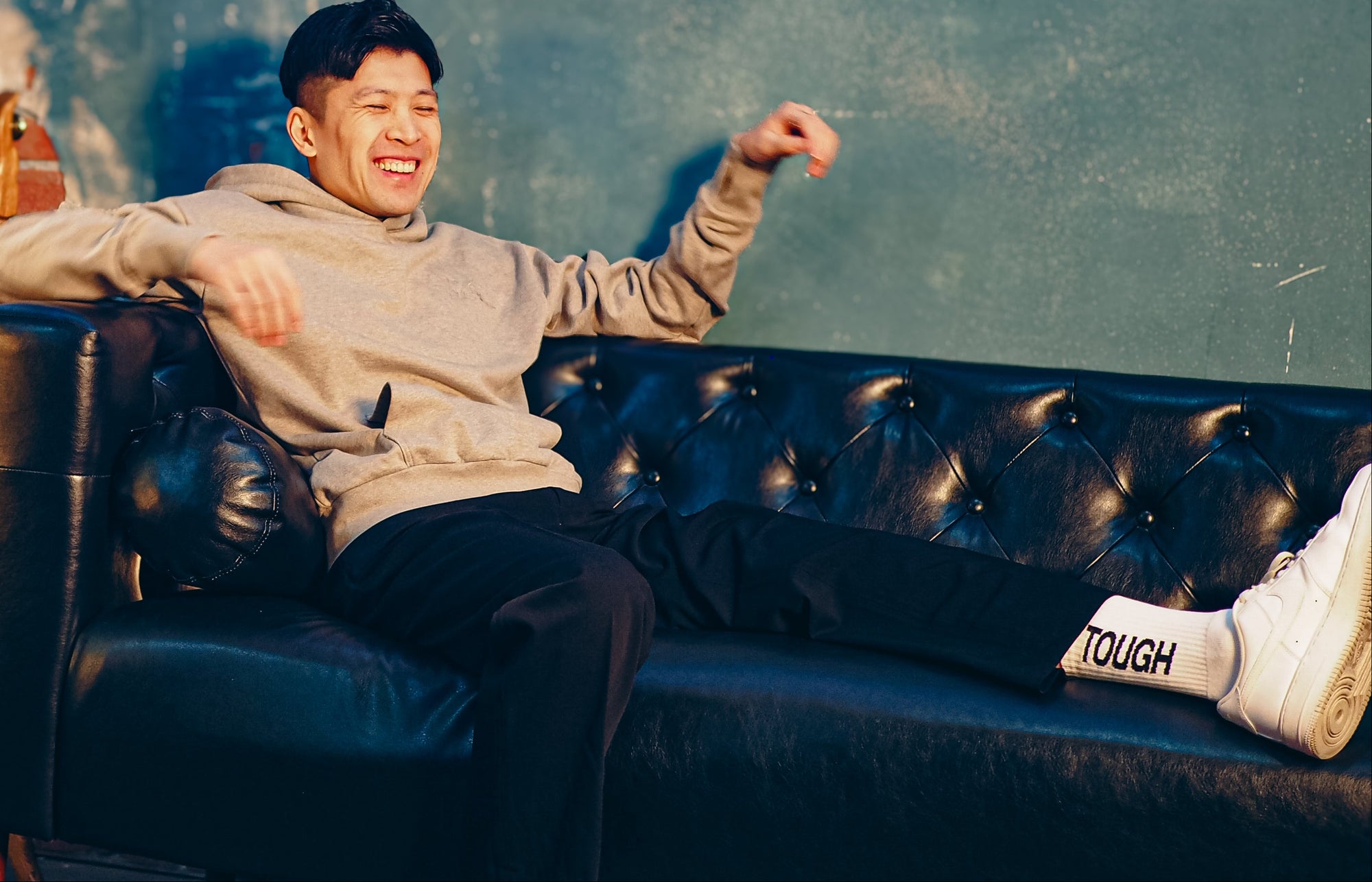 Man sitting on a black leather couch with a mirror and books in the background, wearing socks that say TOUGH on them